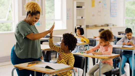 A teacher and student smiling and high-fiving each other in an elementary school classroom setting.